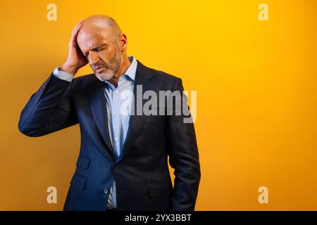 Ein Mann in einem Anzug mit den Händen auf dem Kopf, der verstört aussieht. Er steht vor einem gelben Hintergrund. Stockfoto