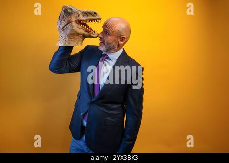 Ein Mann, der eine Dinosauriermaske in der Hand auf gelbem Hintergrund trägt. Stockfoto