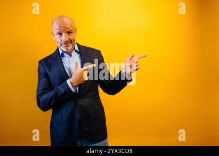 Ein Mann in einem dunkelblauen Anzug zeigt nach rechts. Er blickt mit einem leichten Lächeln in die Kamera. Der Hintergrund ist durchgehend gelb. Stockfoto