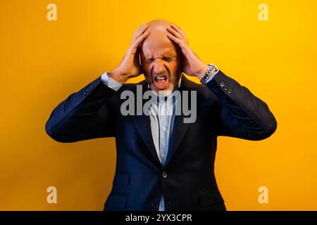 Ein Mann in einem Anzug mit den Händen auf dem Kopf, der verstört aussieht. Er steht vor einem gelben Hintergrund. Stockfoto