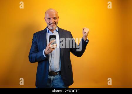 Ein Mann in einem Anzug vor einem gelben Hintergrund. Er hält ein schwarzes Telefon in der rechten Hand und hebt seine linke Hand in die Luft. Er lächelt und sieht sich an Stockfoto