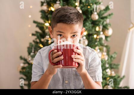 Junge trinkt heiße Schokolade vor dem weihnachtsbaum Stockfoto