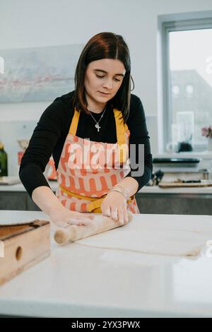 Eine Frau in einer schwarzen Schürze rollt auf einer Theke Teig aus. Sie trägt eine gelbe Schürze mit einem Kreuz drauf Stockfoto