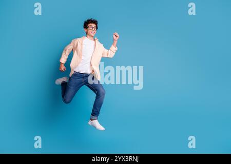 Fröhlicher junger Mann, der begeistert vor einem leuchtend blauen Hintergrund springt. Lässig und stilvoll in trendiger Kleidung. Stockfoto