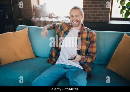 Fröhlicher junger Mann in ungezwungenem Stil, der sich auf einem gemütlichen Sofa in einem hellen Wohnzimmer mit Fernbedienung entspannt Stockfoto