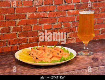 Ein Teller frisch gekochter Garnelen auf einem Salatblatt mit reifer Zitronenscheibe und einem hohen Glas helles Bier auf einem dunklen Holztisch. Nahaufnahme. Stockfoto