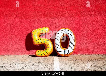 Einige Folien zahlenförmige Ballons in verschiedenen Farben bilden die Zahl 50 auf dem Asphalt, der an einer roten Wand angelehnt ist Stockfoto