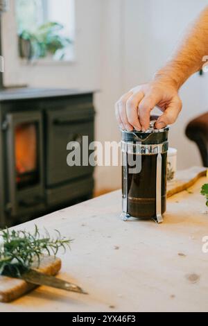 Ein Mann macht Kaffee in einer Kaffeepresse. Die Kaffeepresse steht auf einem Holztisch neben einem Kamin Stockfoto