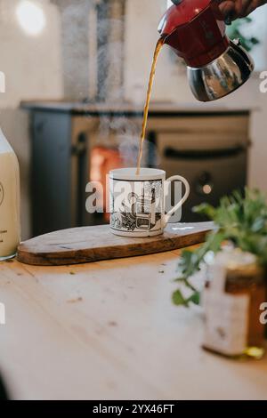 Eine Person gießt Kaffee in eine weiße Tasse mit schwarzem Design. Der Kaffee wird aus einer roten Kaffeekanne gegossen. Die Szene spielt in einer Küche mit Stockfoto