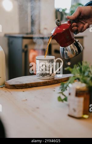 Eine Person gießt Kaffee aus einer roten Kaffeekanne in eine Tasse. Die Kaffeekanne steht auf einem Holztisch neben einem Glas Honig. Die Szene ist gemütlich und einladend Stockfoto