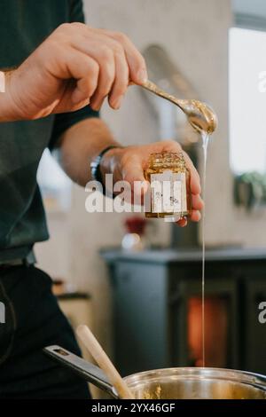 Ein Mann gießt Honig in ein Glas. Der Honig fließt aus dem Glas und auf den Löffel. Der Löffel wird benutzt, um den Honig in das Glas zu schaufeln Stockfoto