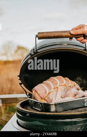 Eine Person kocht ein großes Stück Fleisch auf einem Grill. Das Fleisch ist von Speck umgeben und hat viel Würze darauf. Der Grill ist offen, und der Mea Stockfoto