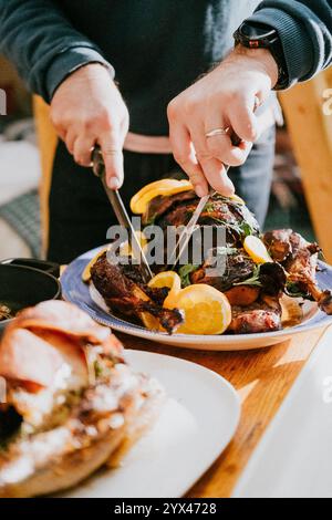 Eine Person schneidet ein gebratenes Huhn mit einem Messer und einer Gabel. Das Huhn wird auf einem blauen Teller mit Orangenscheiben serviert Stockfoto