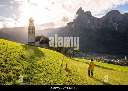 Frau in gelbem Regenmantel genießt Sonnenuntergang über den Dolomiten und der alten Steinkirche in Südtirol, Italien. Stockfoto
