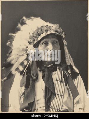 Schlafender Bär, Sioux, 1898. 3/4 Porträt eines Mannes der amerikanischen Ureinwohner, der nach links in Federkopfschmuck schaut, Bandana um den Hals, Holzperlen-Schärpe über die Brust. Stockfoto