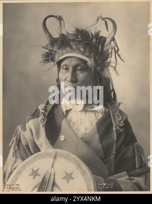 Chief Wets-it der Assiniboine Nation, 1898. Studio-Porträt eines Ureinwohner-Mannes in gehörntem Kopfschmuck, umwickelt in eine Decke, auf die ein Knopf gesteckt ist; er hält einen Schild mit Sternen vor sich. Stockfoto