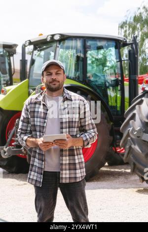 Kaukasischer Mann, der ein digitales Tablet auf dem Hintergrund von Traktoren im Landmaschinenhändler hält. Stockfoto