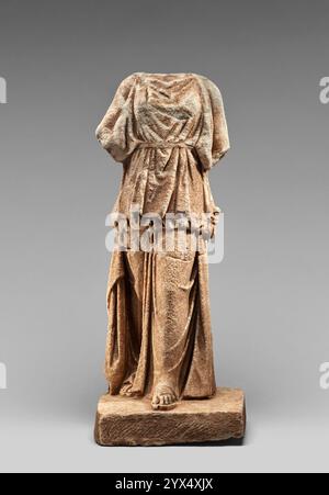 Statuette eines Kore, 350-300 v. Chr. Zusätzliche Info: Diese Statuette einer weiblichen Figur trägt ein Peplos, ein gegurtetes Kleidungsstück mit einer langen Überfalte, die an den Schultern befestigt ist. Sowohl die Figur als auch die quadratische Basis, auf der sie steht, sind aus einem Stück Stein geschnitzt. Der Kopf und die meisten Arme fehlen, aber der Stummel einer Strebe, die mit dem linken Arm verbunden ist, wird auf der linken Hüfte positioniert. Der Oberkörper dreht sich leicht nach links, während das linke Bein nach vorne und das rechte nach hinten gezogen wird. Die Beine werden rechts in der Mitte auf der Basis platziert, wodurch der Körper eine dynamische Qualität erhält, als ob er in erfasst würde Stockfoto