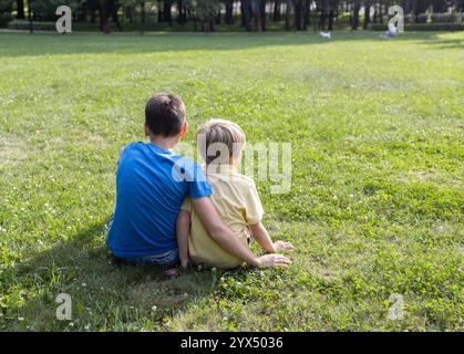 Zwei junge Freunde oder Brüder unterschiedlichen Alters sitzen am sonnigen Sommertag auf dem Rasen im Park und unterhalten sich. Freundschaftskonzept. Rückansicht. Aktive Wiederholung Stockfoto