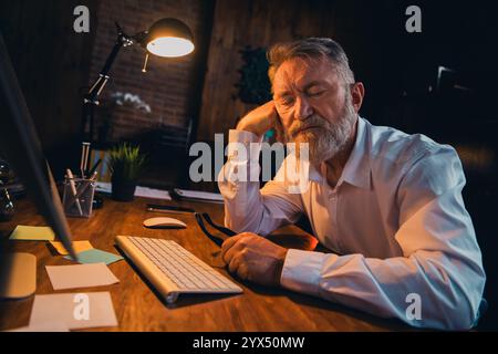 Foto von gutaussehendem reifem Mann müde Schlafbekleidung weißes Hemd formalwear modernes Büro Abendlicht Raum innen Arbeitsplatz Stockfoto