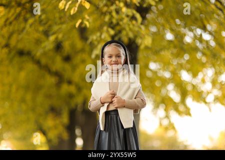 Kindertag. Fröhliches kleines Mädchen 5-6 Jahre alt posiert und lächelt draußen vor der Kamera und steht am Baum im Herbstpark. Stilvoller Zartpressdruck Stockfoto