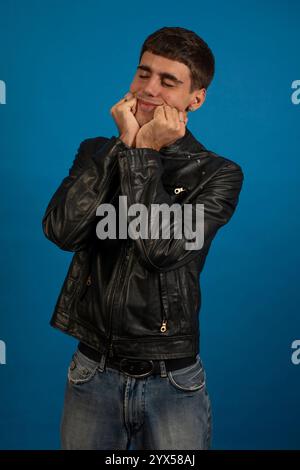 Porträt eines stilvollen jungen Mannes, der sich glücklich und sorglos fühlt, in einer schwarzen Lederjacke und Jeans vor einem leuchtend blauen Hintergrund posiert Stockfoto