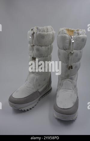 Dinge, Winterstiefel für Damen mit silbernen Schneeflocken und grauen Ledereinsätzen. Schöne Stiefel aus weißem Stoff mit Keilabsatz sind gegen einen weißen Kunststoff gestellt. Stockfoto