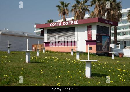 Geschlossener costa Coffee Kiosk, umgeben von Sonnenschirmbasen auf einem grasbewachsenen Gebiet mit gelben Blumen, Palmen und einem Hotel im Hintergrund, das sich auf die Th vorbereitet Stockfoto