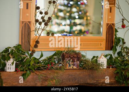 Eine Krippe auf einem Kaminsims. Feenlichter und dekoratives Laub mit einem spiegelreflektierenden Baum. Weihnachtsdekoration in einem Haus in England, Großbritannien. Stockfoto