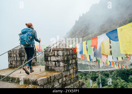 Junge weibliche Trekkerin überquert den Fluss über die Hängebrücke, die mit bunten tibetischen Gebetsfahnen geschmückt ist, die über der Schlucht in der Nähe von Lukla hängen. Everest Base Stockfoto