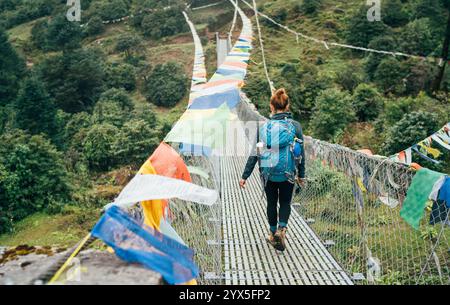 Junge weibliche Trekkerin überquert den Fluss über die Hängebrücke, die mit bunten tibetischen Gebetsfahnen geschmückt ist, die über der Schlucht in der Nähe von Lukla hängen. Everest Base Stockfoto