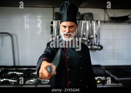 Ein aggressiver Koch mit Bart hält ein Messer in einer Restaurantküche. Er trägt eine schwarze Küchenjacke und einen Hut Stockfoto