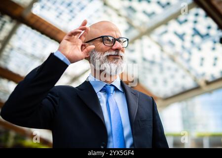 Selbstbewusster Geschäftsmann, der im Freien Haare friert, in scharfem Anzug mit gepflegtem Bart lächelt, moderner Stadtkulisse Stockfoto