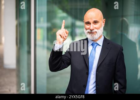 Ein leitender Geschäftsmann in einem scharfen Anzug lächelt und hebt selbstbewusst seinen Zeigefinger, was eine neue Idee in einem modernen Bürogebäude signalisiert Stockfoto