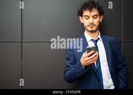 Ein seriöser Geschäftsmann im Anzug liest einen Text vor einem modernen Bürogebäude und konzentriert sich dabei auf sein Smartphone im städtischen Stadtbild Stockfoto