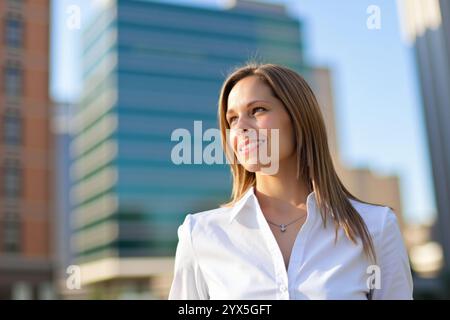 Glückliche Geschäftsfrau, die lächelnd in einer modernen Stadt aufblickt, sich optimistisch und selbstbewusst fühlt Stockfoto