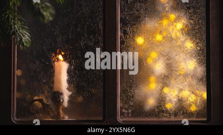 Brennende Kerze sitzt auf einer Fensterbank neben einer mattierten Fensterscheibe mit einem weihnachtsbaum aus Lichterketten, der während eines kalten Winters dahinter leuchtet Stockfoto