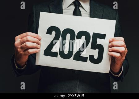 Geschäftsmann, der Papier mit der Nummer 2025 hält, eleganter gutaussehender Unternehmer im dunklen Interieur, Neujahrskonzept, selektiver Fokus Stockfoto