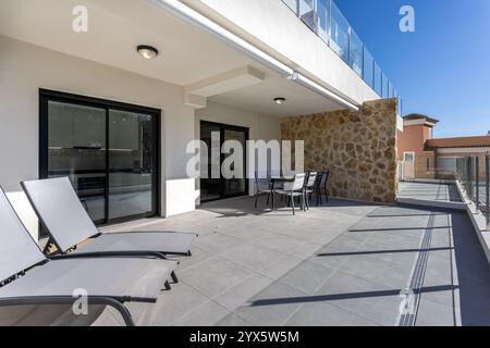 Moderne Terrasse mit Liegestühlen, einem Essgarnitur im Freien, Glasgeländern und einer Wand mit Steinakzenten, perfekt zum Entspannen oder Essen im Freien. Stockfoto