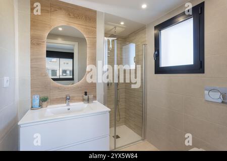 Modernes Bad mit bodenebener Dusche, holzstrukturierten Details, beleuchtetem ovalem Spiegel, Milchfenster und elegantem weißen Kosmetikbereich. Stockfoto