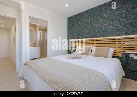 Modernes Schlafzimmer mit auffälligen geblümten Tapeten, Holzkopfteil, gemütlicher Bettwäsche und eigenem Bad mit minimalistischem Design. Stockfoto