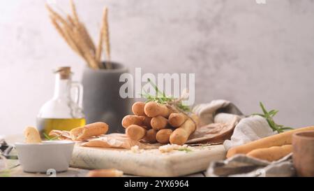 Rustikale Brotstangen mit Garn gebunden und mit Rosmarin auf einem Holzbrett garniert, mit Olivenöl und Weizenstielen im Hintergrund, die ein gemütliches k schaffen Stockfoto