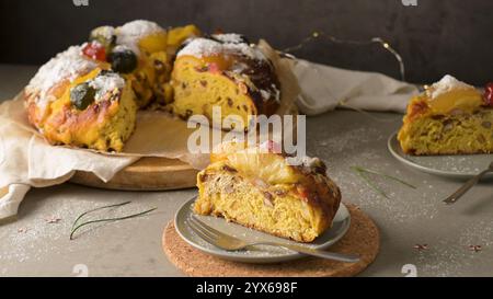 Festlicher Fruchtkuchen mit Puderzucker, geschmückt mit bunten kandierten Früchten, serviert auf einem Holzbrett mit einer Scheibe auf einem Teller, bereit zum Genießen Stockfoto