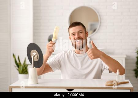 Glücklicher junger Mann mit Haarausfall, der den Kamm hält und zu Hause den Daumen nach oben zeigt Stockfoto