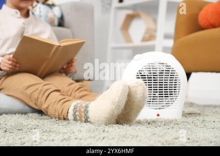 Netter Junge in warmen Socken mit elektrischer Heizlüfter, der Buch auf Teppich zu Hause liest, Großaufnahme Stockfoto