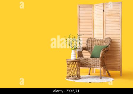 Tisch mit Bambusstämmen in Vase, Korbsessel und Klappschirm auf gelbem Hintergrund Stockfoto