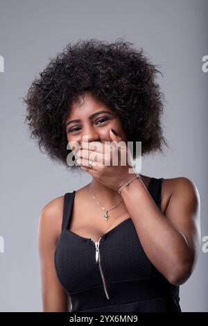 Studio-Porträt einer fröhlichen jungen Frau mit lockigen Haaren, die ein schwarzes Top trägt, ihren Mund mit der Hand bedeckt, während sie über einen grauen Hintergras lacht Stockfoto