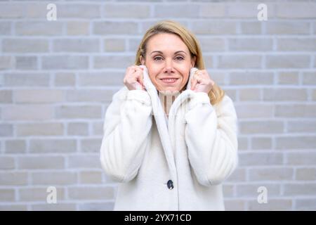 Eine blonde Frau in einem weichen weißen Mantel lächelt warm, während sie sanft ihren Kragen hält. Stockfoto