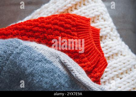 Graue, weiße und leuchtend orange Strickpullover auf einem Stapel. Ein Stapel kuscheliger, handgefertigter Kleidung. Konzept für unterschiedliche Strickmuster. Farbenfrohe gewebte Pullover Stockfoto