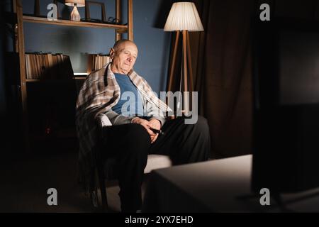 Einsamer Großvater Rentner, in eine Decke gewickelt, schläft auf einem Stuhl im Wohnzimmer und schaut nachts fern müder alter Mann Stockfoto